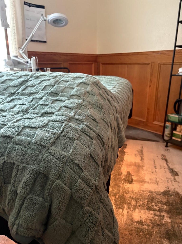 Peaceful treatment room with facial bed, warm ambiance, and pro skincare tools ready for client treatments. This is a cozy space with a weighted blanket to help relaxation and destressing while enjoying a professional service.